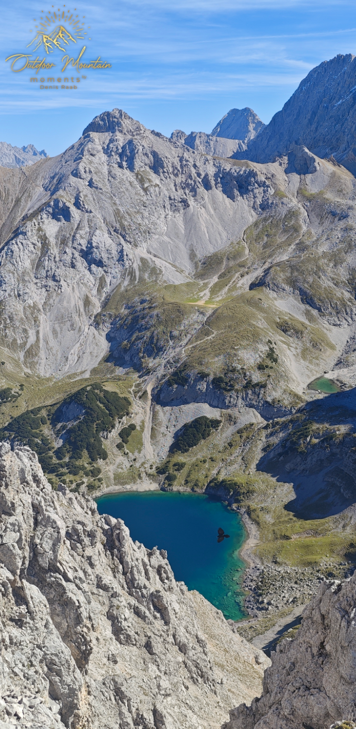 Blick auf einen Gebirgssee umgeben von steilen Felsen und grünen Wiesen.