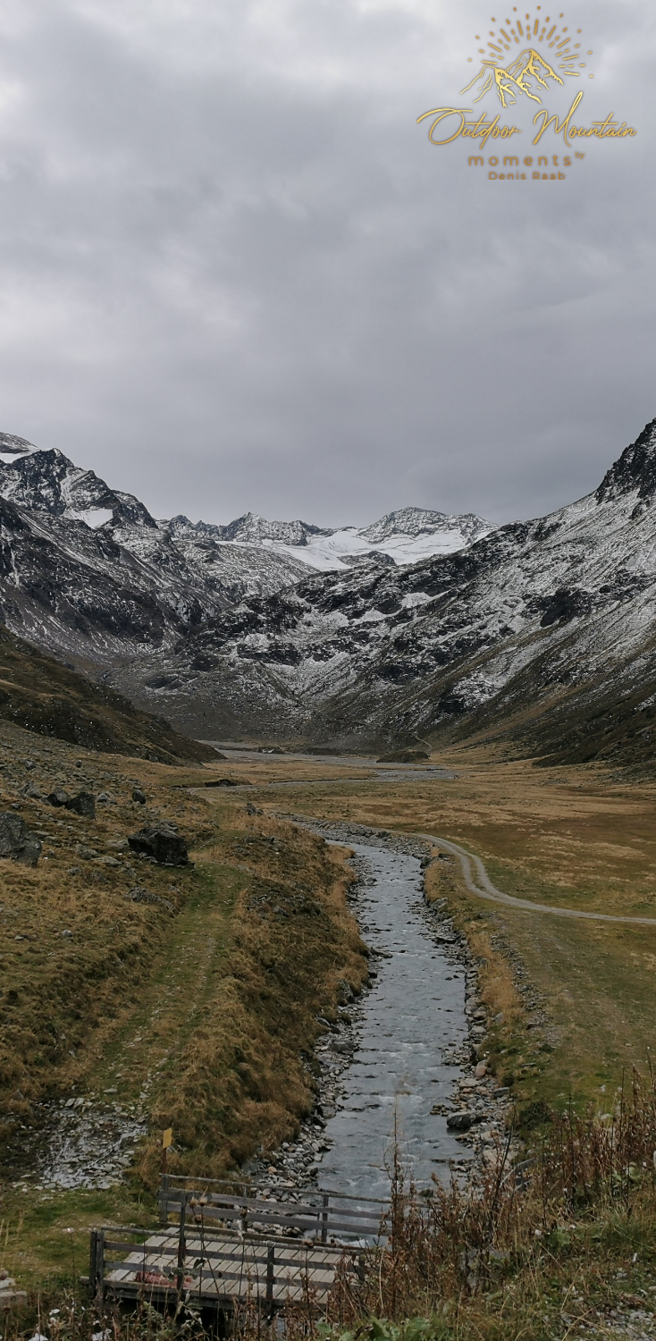 Berglandschaft mit einem Fluss, grünen Wiesen und schneebedeckten Gipfeln.